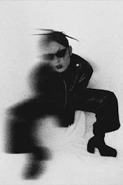 A person dressed in dark, avant-garde clothing is partially obscured by a blur effect. The person is wearing oversized sunglasses and has their hair styled upward. The background appears to be a plain, light-colored surface. The overall image quality is grainy, giving it a vintage or artistic feel.