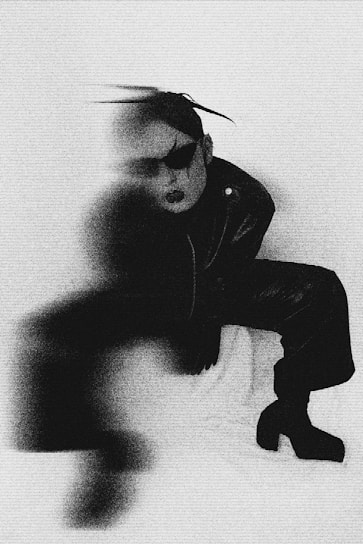 A person dressed in dark, avant-garde clothing is partially obscured by a blur effect. The person is wearing oversized sunglasses and has their hair styled upward. The background appears to be a plain, light-colored surface. The overall image quality is grainy, giving it a vintage or artistic feel.