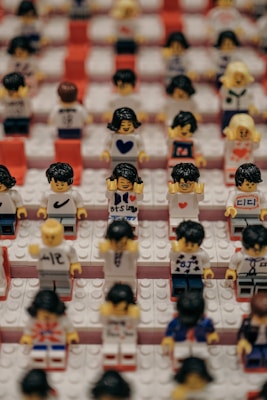 Rows of Lego figurines are arranged neatly on a platform with red and white brick patterns. Each figurine appears unique, showcasing different facial expressions and attire, some with printed messages or symbols on their shirts, such as hearts and letters.
