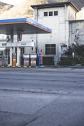 An empty gas station is situated in front of an old industrial building with a slightly worn appearance. The gas station has multiple fueling pumps under a large canopy with blue and orange accents. The surrounding area includes some greenery and trees on a hilly landscape in the background.