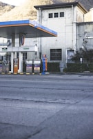 An empty gas station is situated in front of an old industrial building with a slightly worn appearance. The gas station has multiple fueling pumps under a large canopy with blue and orange accents. The surrounding area includes some greenery and trees on a hilly landscape in the background.