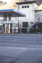 An empty gas station is situated in front of an old industrial building with a slightly worn appearance. The gas station has multiple fueling pumps under a large canopy with blue and orange accents. The surrounding area includes some greenery and trees on a hilly landscape in the background.