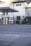 An empty gas station is situated in front of an old industrial building with a slightly worn appearance. The gas station has multiple fueling pumps under a large canopy with blue and orange accents. The surrounding area includes some greenery and trees on a hilly landscape in the background.