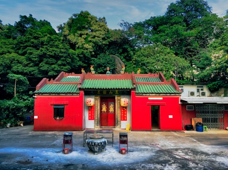 A small traditional temple with red walls and a green tiled roof is surrounded by lush green trees. The temple features ornate decorative elements and has an incense burner placed at the front. The setting is peaceful and isolated, with a concrete courtyard leading up to the entrance.