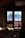 A cozy wooden cabin interior with a window view of snow-covered mountains. Sunlight spills through the window, illuminating a bed with rumpled sheets. The curtains frame the view and the warm brown tones of the wood contrast with the bright snow outside.