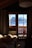 A cozy wooden cabin interior with a window view of snow-covered mountains. Sunlight spills through the window, illuminating a bed with rumpled sheets. The curtains frame the view and the warm brown tones of the wood contrast with the bright snow outside.