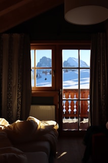 A cozy wooden cabin interior with a window view of snow-covered mountains. Sunlight spills through the window, illuminating a bed with rumpled sheets. The curtains frame the view and the warm brown tones of the wood contrast with the bright snow outside.