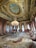 A luxurious ornate room with an intricate, gold-accented ceiling featuring a central fresco. The room is furnished with red and gold upholstered chairs and sofas, a large central chandelier, and an elaborate patterned carpet.