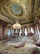 A luxurious ornate room with an intricate, gold-accented ceiling featuring a central fresco. The room is furnished with red and gold upholstered chairs and sofas, a large central chandelier, and an elaborate patterned carpet.