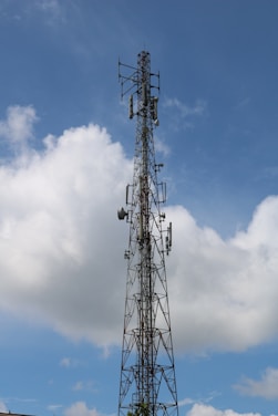 A tall metal telecommunications tower extends into the sky, surrounded by white clouds. The structure includes several antennas and dishes, indicating its use for broadcasting or communication.