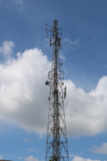 A tall metal telecommunications tower extends into the sky, surrounded by white clouds. The structure includes several antennas and dishes, indicating its use for broadcasting or communication.