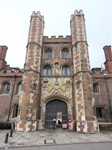 A grand entrance with two large, ornate brick towers flanking a central archway. The architecture features detailed carvings and a coat of arms above the wooden doors. The stone pavement leads to the entrance, and a sign is visible near the door.