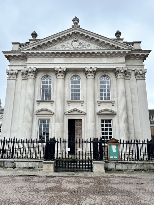 A classical building features tall, ornate columns and large, arched windows, topped with an intricately carved pediment. The entrance is adorned with a heavy wooden door, and the structure is surrounded by a black wrought iron fence.