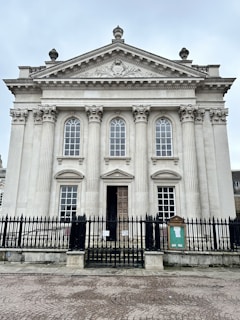 A classical building features tall, ornate columns and large, arched windows, topped with an intricately carved pediment. The entrance is adorned with a heavy wooden door, and the structure is surrounded by a black wrought iron fence.
