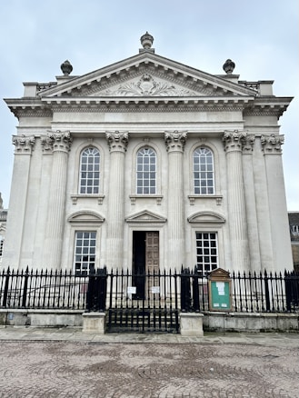 A classical building features tall, ornate columns and large, arched windows, topped with an intricately carved pediment. The entrance is adorned with a heavy wooden door, and the structure is surrounded by a black wrought iron fence.