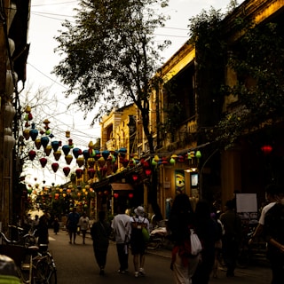 A bustling street in an old town features hanging lanterns in various colors strung across the road. People are walking along the street, surrounded by historic buildings with a weathered charm. The scene is illuminated by a warm, golden light, creating a lively yet relaxed atmosphere.