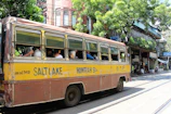 A vintage yellow and brown bus, labeled with Salt Lake and Howrah, is traveling down a street. Several passengers are seated inside, with the windows open. The background features a row of old buildings with greenery and shops. The scene is lively with people walking and street activity.