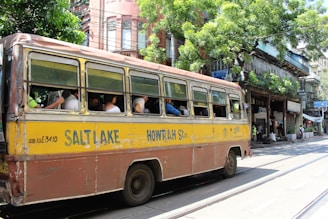 A vintage yellow and brown bus, labeled with Salt Lake and Howrah, is traveling down a street. Several passengers are seated inside, with the windows open. The background features a row of old buildings with greenery and shops. The scene is lively with people walking and street activity.