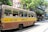 A vintage yellow and brown bus, labeled with Salt Lake and Howrah, is traveling down a street. Several passengers are seated inside, with the windows open. The background features a row of old buildings with greenery and shops. The scene is lively with people walking and street activity.