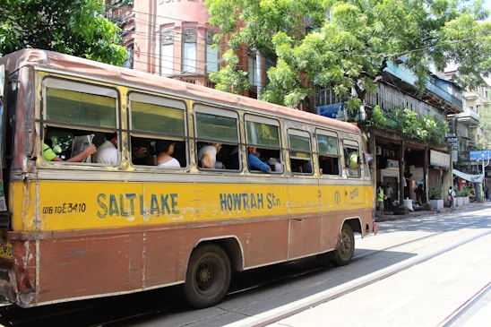 A vintage yellow and brown bus, labeled with Salt Lake and Howrah, is traveling down a street. Several passengers are seated inside, with the windows open. The background features a row of old buildings with greenery and shops. The scene is lively with people walking and street activity.