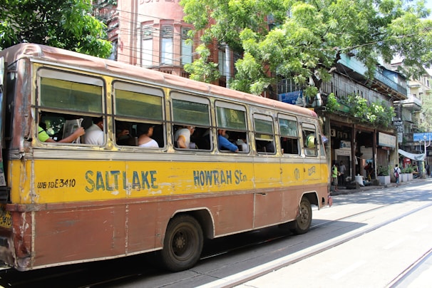 A vintage yellow and brown bus, labeled with Salt Lake and Howrah, is traveling down a street. Several passengers are seated inside, with the windows open. The background features a row of old buildings with greenery and shops. The scene is lively with people walking and street activity.