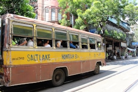 A vintage yellow and brown bus, labeled with Salt Lake and Howrah, is traveling down a street. Several passengers are seated inside, with the windows open. The background features a row of old buildings with greenery and shops. The scene is lively with people walking and street activity.
