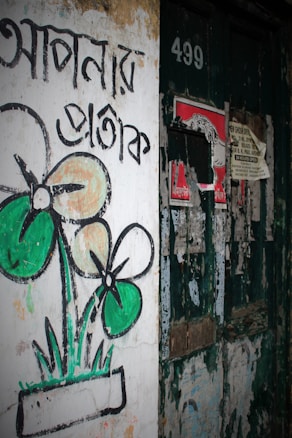 An aged wall with faded paint, featuring graffiti of a stylized flower in a pot with Bengali script beside it. The wall shows signs of wear with peeling paint and old, tattered posters nearby. The number 499 is visible on the door.