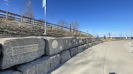 A pathway beside a stone wall displaying plaques and an inscription about indigenous history. The sky is clear and blue, with a wooden fence and leafless trees in the background. Flags and distant structures are visible towards the end of the path.
