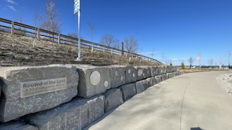 A pathway beside a stone wall displaying plaques and an inscription about indigenous history. The sky is clear and blue, with a wooden fence and leafless trees in the background. Flags and distant structures are visible towards the end of the path.