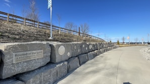 A pathway beside a stone wall displaying plaques and an inscription about indigenous history. The sky is clear and blue, with a wooden fence and leafless trees in the background. Flags and distant structures are visible towards the end of the path.