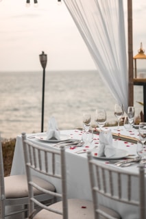 A neatly arranged table with white linens is set for an event beside a body of water. The table is adorned with glassware, plates with white napkins folded elegantly, and scattered red rose petals. A sheer white drape is suspended as part of the decor, and a lantern and torch are present in the background. The setting suggests a peaceful, elegant outdoor atmosphere.