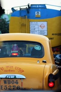 A yellow taxi is seen in traffic with a blue and yellow vehicle in front of it. The taxi has a registration plate and religious stickers on the rear window. There is text in a regional script on the back of the taxi.