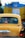 A yellow taxi is seen in traffic with a blue and yellow vehicle in front of it. The taxi has a registration plate and religious stickers on the rear window. There is text in a regional script on the back of the taxi.