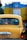 A yellow taxi is seen in traffic with a blue and yellow vehicle in front of it. The taxi has a registration plate and religious stickers on the rear window. There is text in a regional script on the back of the taxi.