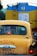 A yellow taxi is seen in traffic with a blue and yellow vehicle in front of it. The taxi has a registration plate and religious stickers on the rear window. There is text in a regional script on the back of the taxi.