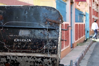 A close-up view of a weathered hand-pulled rickshaw in a narrow urban street. The rickshaw's surface shows rust and peeling paint with the word 'Kolkata' stenciled onto it. In the background, there are vibrantly painted walls in shades of blue and red. A person in a white shirt is walking away in the distance.
