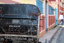 A close-up view of a weathered hand-pulled rickshaw in a narrow urban street. The rickshaw's surface shows rust and peeling paint with the word 'Kolkata' stenciled onto it. In the background, there are vibrantly painted walls in shades of blue and red. A person in a white shirt is walking away in the distance.