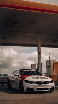 A sleek, sporty car with racing decals is parked at a gas station. The car is adorned with branding such as Marlboro and Toyo Tires, and features the number 82. In the background, there is an overcast sky and another vehicle partially visible. The gas station canopy and pumps are prominently shown.