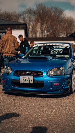 A vibrant blue sports car with customized features, including a prominent hood scoop and distinctive front bumper, parked in an outdoor setting. Several people are gathered nearby, some appearing to admire the vehicle. The car has personalized license plates and stickers, emphasizing its modified racing style.