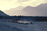 A convoy of Lambretta riders cruising through a forested mountain route under clear blue skies.