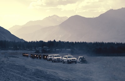 A convoy of motorcycles followed by cars travels through a dusty landscape with a backdrop of imposing mountains. The sky is overcast with a mix of clouds and sunlight, casting muted shadows on the terrain. A row of trees lines the base of the mountains, adding to the sense of depth and scale.
