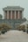 A large, gray stone mausoleum structure features tall pillars and a tiered roof. The pathway leading up to the building is lined with carefully pruned potted trees. The overall scene has a symmetrical and formal appearance, with a subdued ambiance.