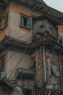 A weathered and deteriorating building facade with a mixture of architectural elements and exposed wiring. The walls exhibit significant wear and peeling paint, while open shutters reveal a lack of maintenance. A large metal tank is attached to the exterior, contributing to the cluttered appearance.