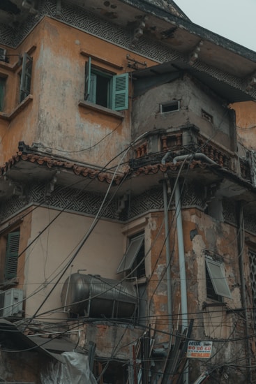 A weathered and deteriorating building facade with a mixture of architectural elements and exposed wiring. The walls exhibit significant wear and peeling paint, while open shutters reveal a lack of maintenance. A large metal tank is attached to the exterior, contributing to the cluttered appearance.
