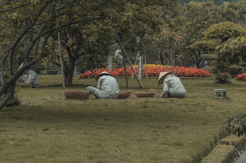 Several people wearing wide-brimmed hats are tending to a garden. They are positioned on a grassy area surrounded by trees and vibrant flowers, with baskets placed beside them. The setting appears peaceful and natural, with a focus on greenery and outdoor work.