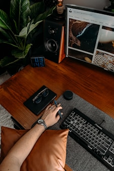 A wooden desk with a keyboard and a monitor displaying a webpage is shown. A person wearing a watch rests their hand on a brown pillow, holding a pen. Nearby are a black speaker, a pair of sunglasses on a notepad, a small digital clock, and a plant.