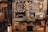An eclectic bakery display at a café features handwritten signs advertising various baked goods and drinks. String lights, a chalkboard menu, and a Nescafé advertisement add charm to the wooden shelves which hold pastries and desserts such as walnut pie, apple almond cake, and brownies. A mixture of rustic and cozy elements with various signs creates an inviting atmosphere.