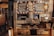 An eclectic bakery display at a café features handwritten signs advertising various baked goods and drinks. String lights, a chalkboard menu, and a Nescafé advertisement add charm to the wooden shelves which hold pastries and desserts such as walnut pie, apple almond cake, and brownies. A mixture of rustic and cozy elements with various signs creates an inviting atmosphere.