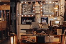 An eclectic bakery display at a caf&eacute; features handwritten signs advertising various baked goods and drinks. String lights, a chalkboard menu, and a Nescaf&eacute; advertisement add charm to the wooden shelves which hold pastries and desserts such as walnut pie, apple almond cake, and brownies. A mixture of rustic and cozy elements with various signs creates an inviting atmosphere.