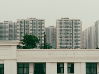 A series of high-rise apartment buildings dominates the skyline, with uniform design and numerous windows. In the foreground, there is a white building with a flat roof and a contrasting green tree. This urban scene suggests dense population and modern city living.
